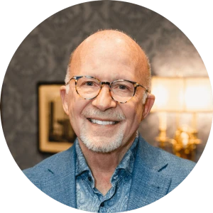 A smiling older man with glasses, blue blazer, light-blue shirt, in a warmly lit room with patterned wallpaper.