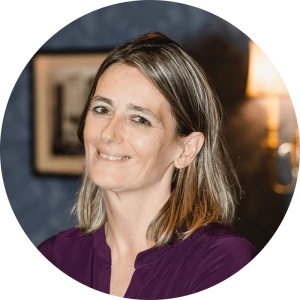 A smiling woman with shoulder-length light brown hair, wearing a purple top in a circular portrait.