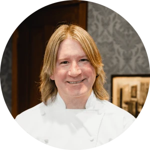 A smiling chef with shoulder-length blond hair wearing a white chef&rsquo;s coat, in a warmly lit kitchen or dining area.