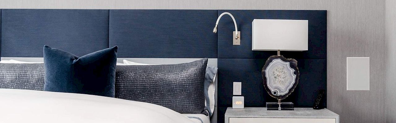 A modern bedroom with a blue headboard, a bed with pillows, and a nightstand holding a lamp, book, and phone.