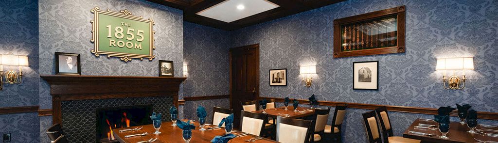 A formal dining room with blue patterned wallpaper, wooden tables set for a meal, blue glassware, and a lit fireplace labeled &ldquo;1855 Room.&rdquo;