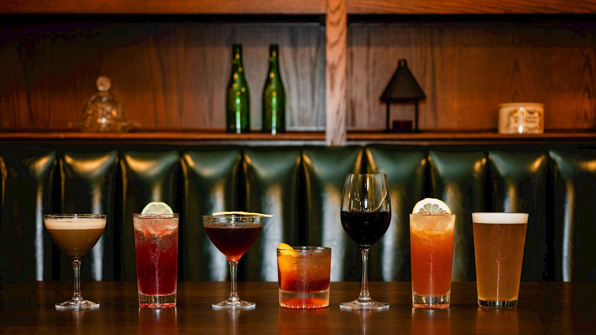 A row of seven cocktails on a bar counter, each with different colors and garnishes, set against a dark green booth and wooden shelves.