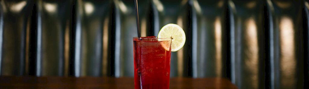 A tall red cocktail with two straws and a lime wheel garnish sits on a wooden bar, backed by dark teal booth seating, in a cozy lounge.