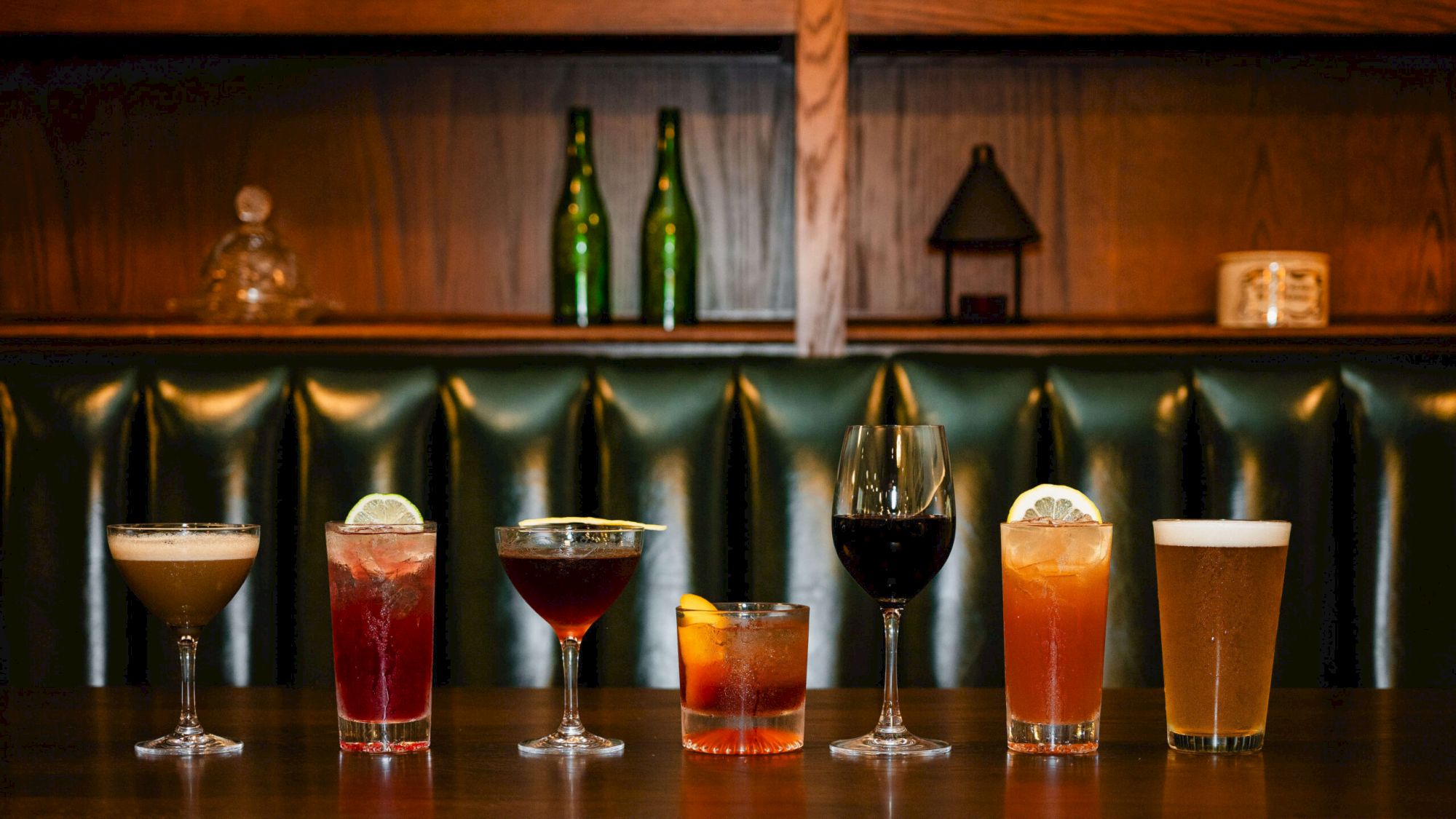 A row of seven assorted cocktails on a bar, each with different colors and garnishes, in front of a green upholstered bar back.