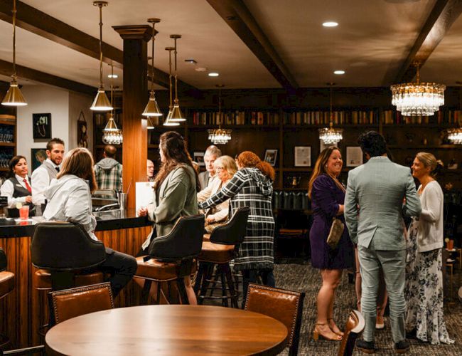 A lively bar scene with people mingling at the counter, chatting in groups, warm lighting, and wooden tables in a cozy lounge atmosphere.