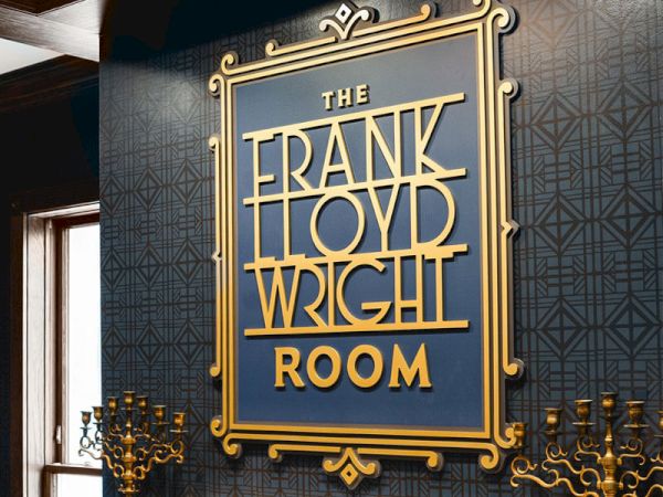 A stylish wall plaque reads &ldquo;The Frank Lloyd Wright Room&rdquo; in gold lettering, framed with ornate detailing, set against a dark patterned backdrop.