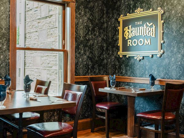 A cozy, vintage-style cafe or pub interior with dark textured walls, wooden tables and chairs, a window, and a framed sign reading &ldquo;bar/haunted room.&rdquo;