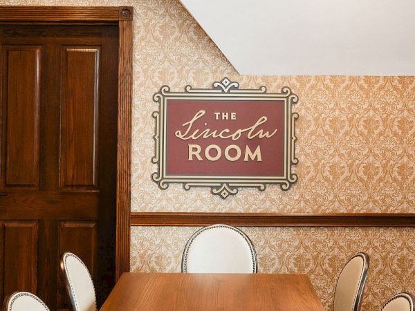 A wooden conference table in a room with vintage wallpaper, a sign reading &ldquo;The Lincoln Room&rdquo; hangs on the wall beside a door, meeting chairs around the table.