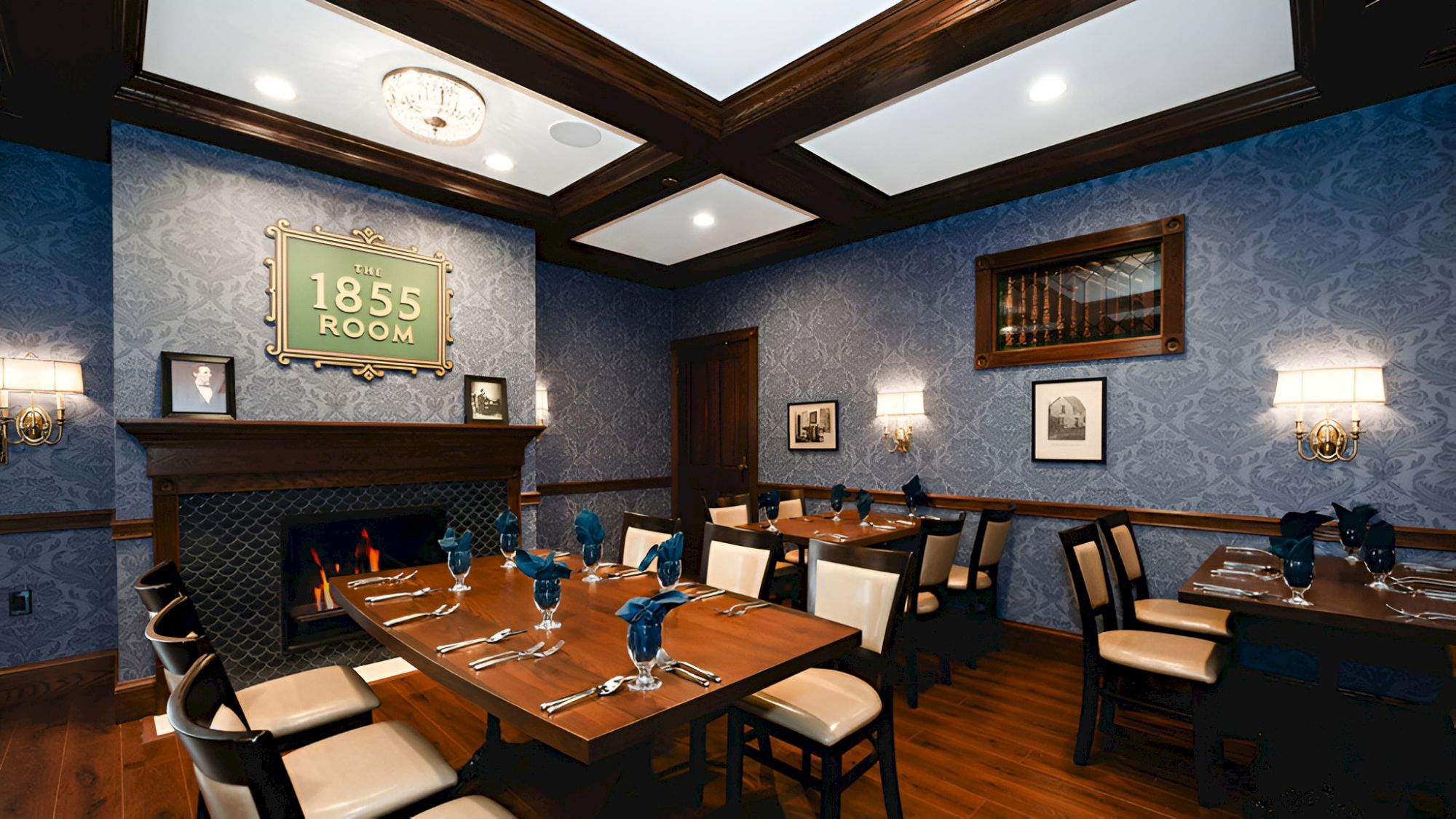 A formal dining room with a long wooden table set for a meal, blue patterned walls, and a cozy fireplace in the background.
