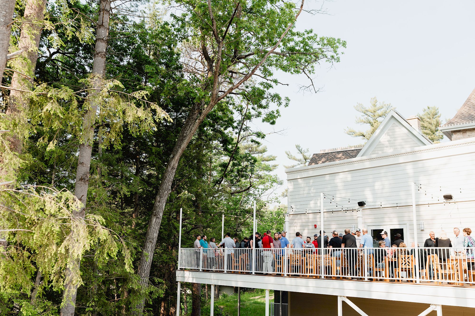 An outdoor event crowd gathers on a deck by a white building, surrounded by tall green trees and summer sky.