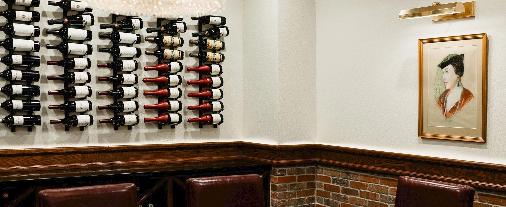 A wine wall with neatly arranged bottles on racks, a brick-haloed lower wall, and a framed artwork alongside seating in a cozy tasting area.