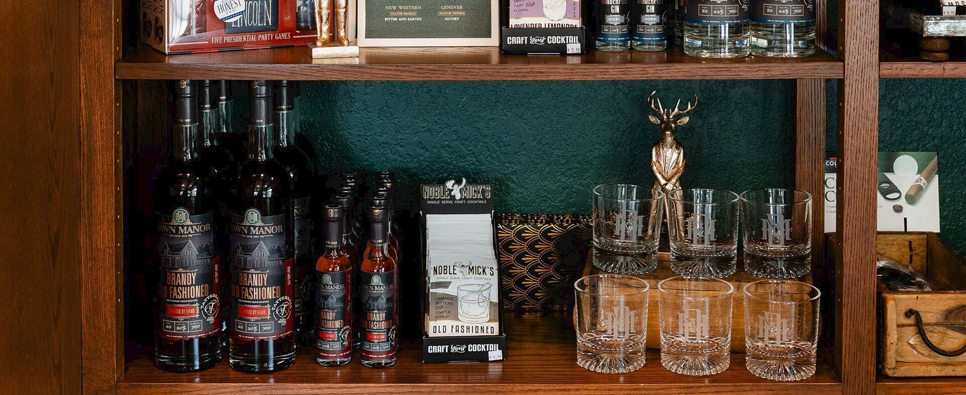 A wooden shelf display with bottles of liquor on the left, a small box, several clear whiskey glasses on the right, and a decorative stag figurine in the center, all against a green wall.