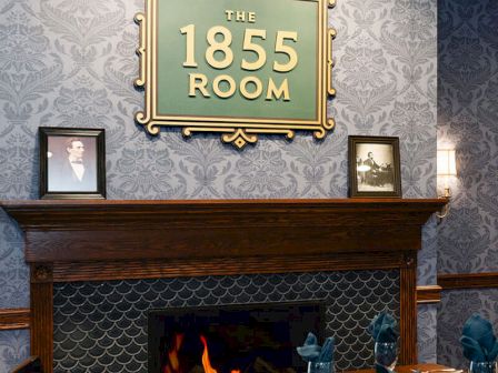 A cozy dining space in a restaurant named &ldquo;The 1855 Room,&rdquo; with a lit fireplace, dark wood mantle, patterned wallpaper, and framed photos above.