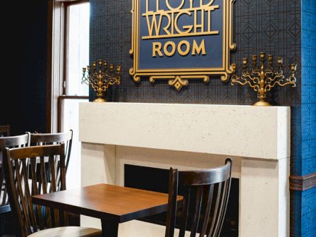 A cozy corner of a Frank Lloyd Wright Room with a fireplace, dark walls, wooden chairs, and a decorative sign above the mantel. Top it at 140 characters, ending with a period.