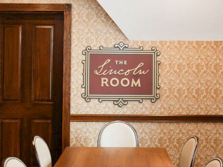 A wooden conference table in a vintage-lounge room, ornate wallpaper, and a sign reading &ldquo;The Lincoln Room&rdquo; on the wall.