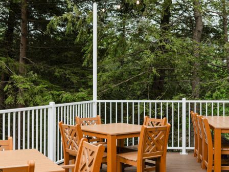 Outdoor wooden deck with multiple square tables and chairs, white railings, and a forest backdrop.