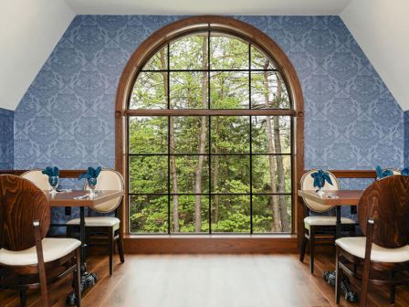 A cozy dining nook with a large arched window, blue patterned walls, wooden chairs, and a view of greenery outside, warm and inviting.