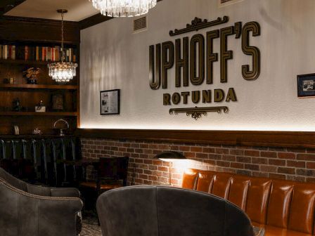 A chic dining area with a brick wall, leather booths, plush chairs, chandeliers, and a bold sign reading "UPNOFE'S ROTA" on the back wall.