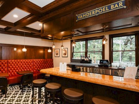 A cozy, vintage bar with a wooden counter, black-and-white tile floor, red upholstered booths, and warm hanging lights at Vanderpoel Bar.