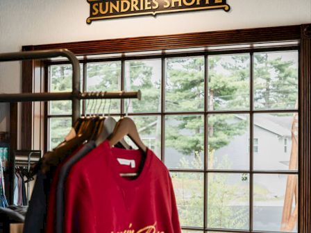A cozy shop interior with hanging clothes on racks and a red sweatshirt in the foreground, under a window view and a vintage sign.