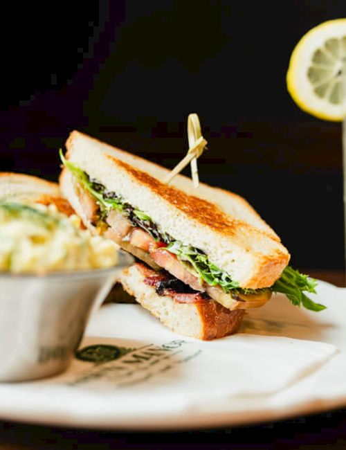 A plated sandwich with greens, a small side dish (perhaps potato salad), and a lemonade with a lemon wheel on a straw; everything on a table.