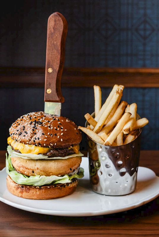 A burger with sesame bun, cheddar, lettuce, and a knife stuck through the top, served with a side of fries on a plate.