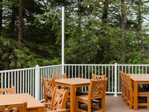 Outdoor wooden patio with several empty tables and chairs, surrounded by a white railing and tall trees, sunny and serene.