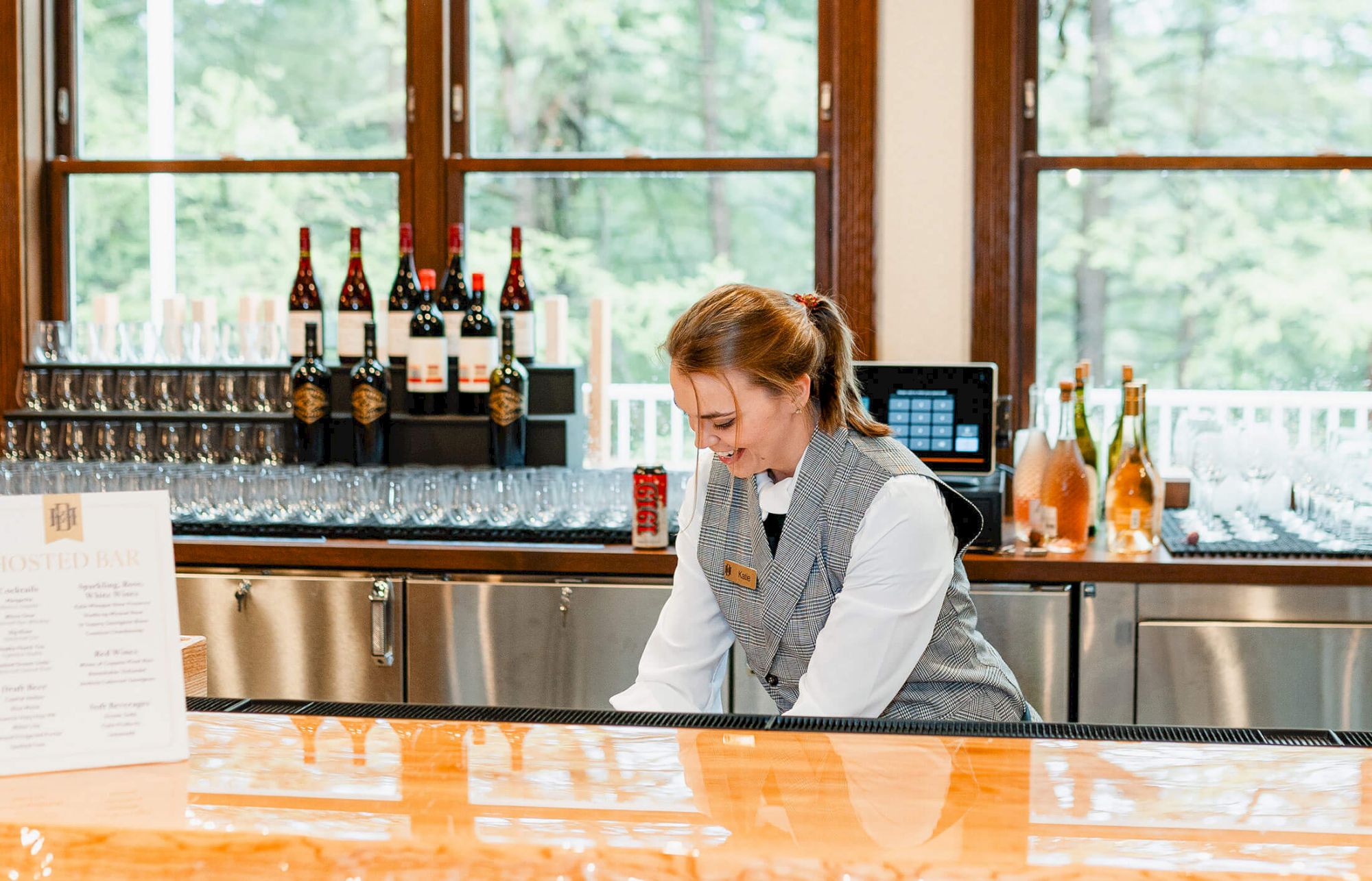 A smiling bartender in a gray vest pours at a polished wooden bar, bottles lined behind, windows and natural light in the background.