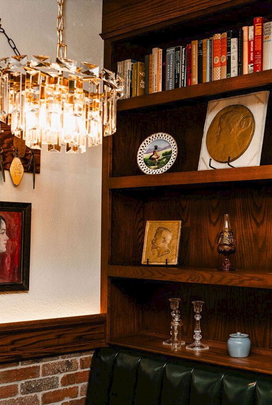 A cozy corner with a brick baseboard, wooden shelves, a chandelier, framed art, and various books and trinkets displayed above a dark tufted bench, inviting warm vibes.