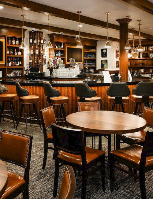 A cozy bar with wooden tables and leather chairs, a long counter, shelves of bottles, and warm ambient lighting.