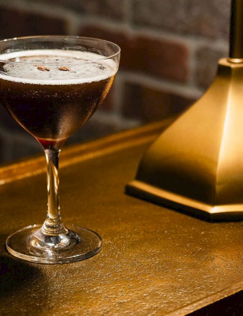 A coupe glass with a dark cocktail beside a brass lamp on a wooden surface, cozy bar vibe.