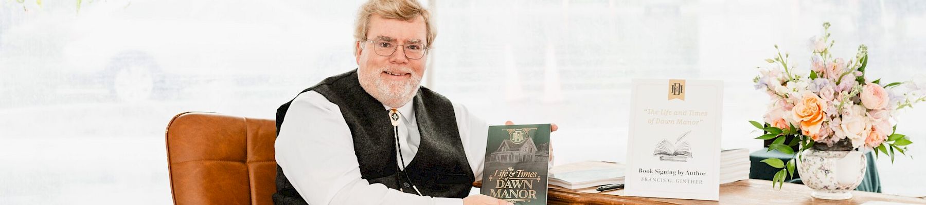 An older man in a vest sits at a wooden desk holding a book, with a flower bouquet and decorative items on the desk.