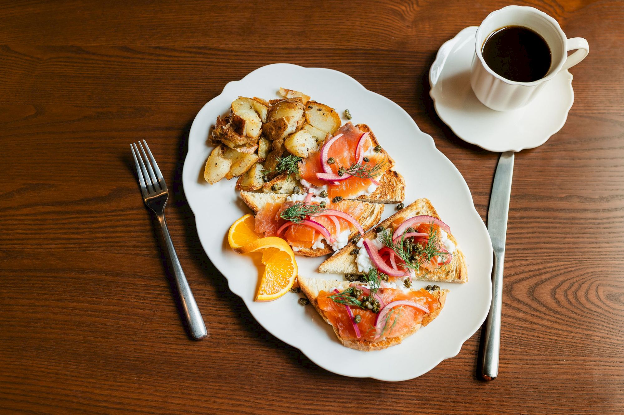 A plate of assorted bruschetta slices with herbs, tomatoes, and citrus garnish, plus crusty bread chunks, a fork, knife, and a cup of coffee on a wooden table.