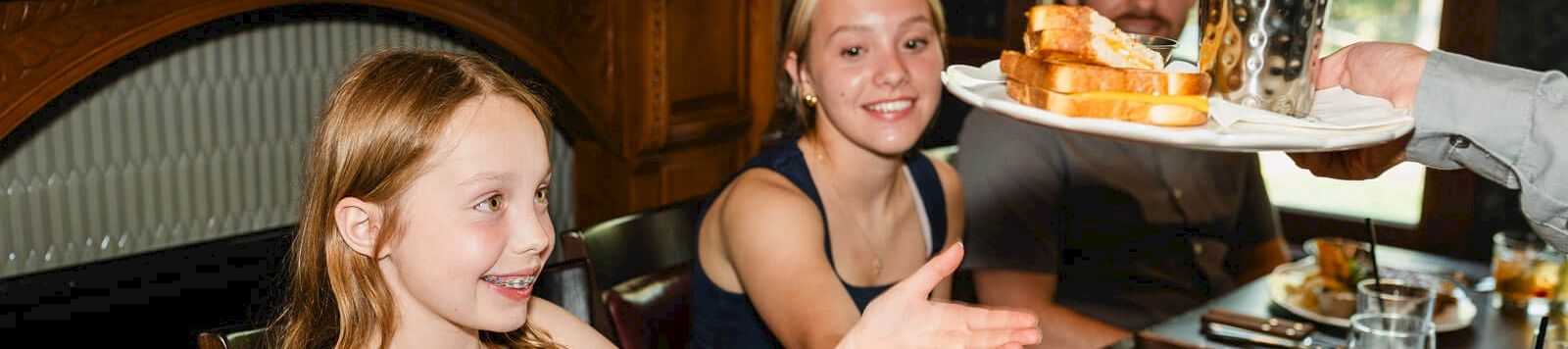 Three people sit at a dining table in a restaurant; a girl in a black dress waves while others smile as a server carries a tray of drinks and food.