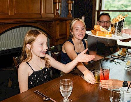 Three people sit at a dining table in a restaurant; a girl in a black dress waves while others smile as a server carries a tray of drinks and food.