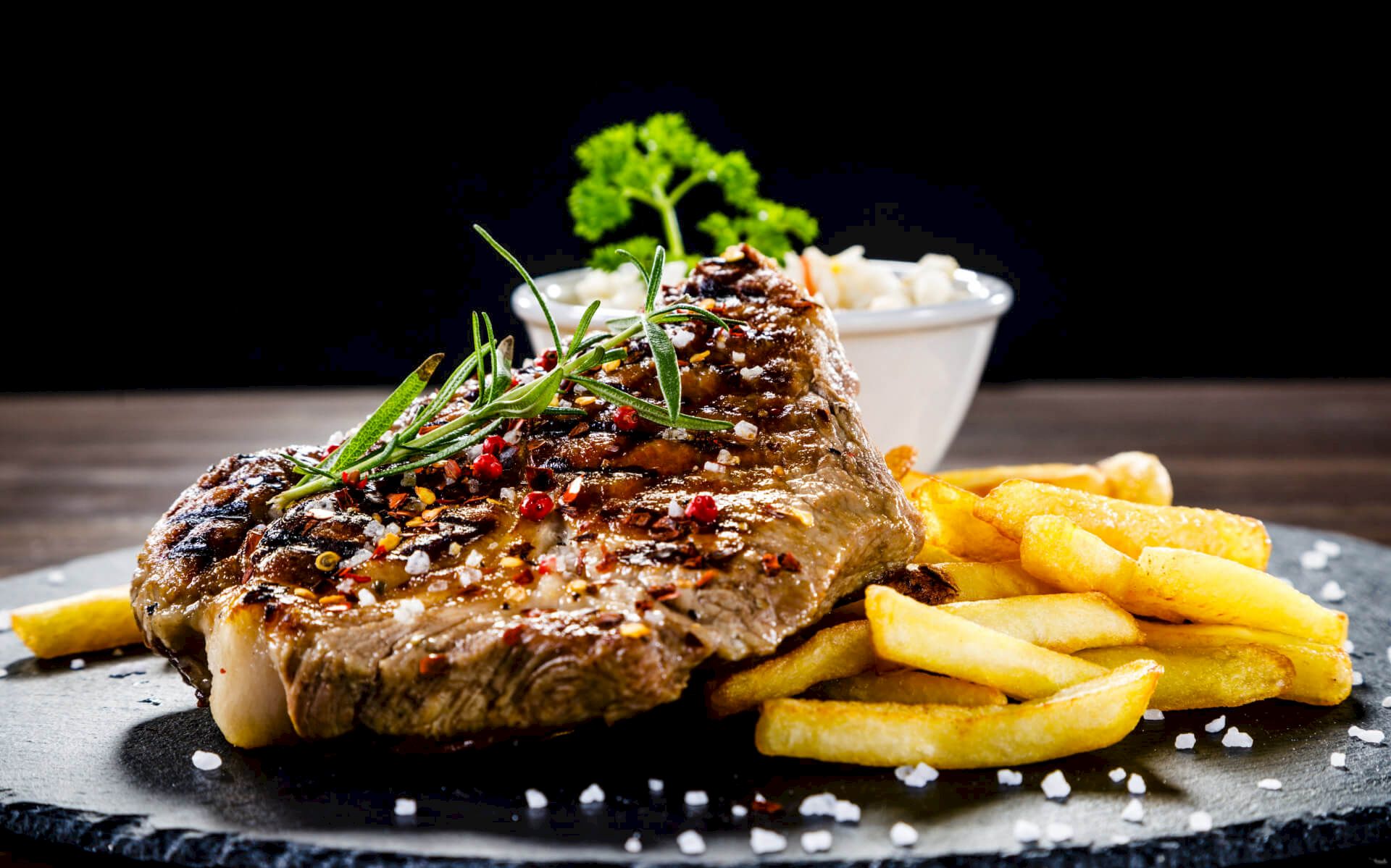A grilled steak with herbs and sesame, served on a dark plate with crispy fries and a small bowl of sauce in the background.