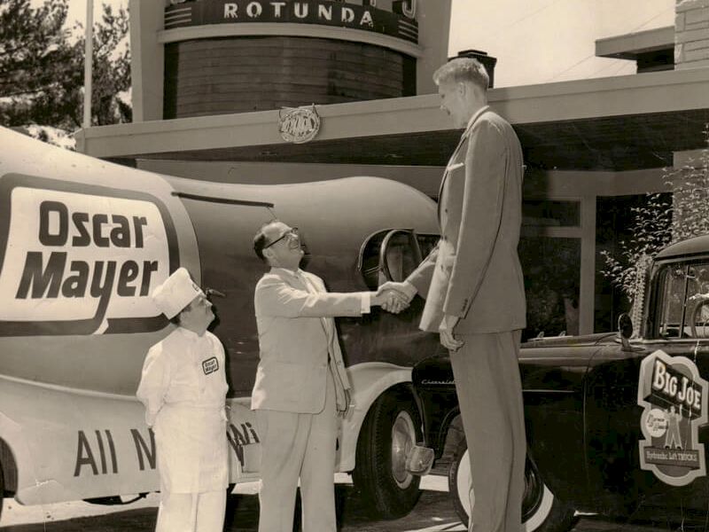 A vintage black-and-white scene with three men shaking hands beside a classic car in front of a theater marquee, &ldquo;Topper&rdquo; style signage, and an &ldquo;Oscar Mayer&rdquo; delivery ad.