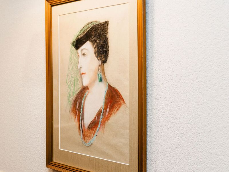 A framed portrait of a woman with a dark bun, wearing a red top and dangling turquoise earrings, hung on a light-textured wall.