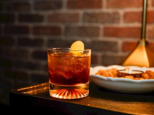 A glass of whiskey on the rocks with a lemon twist sits beside a plate of fried or breaded snacks on a dark wooden table under warm lighting.