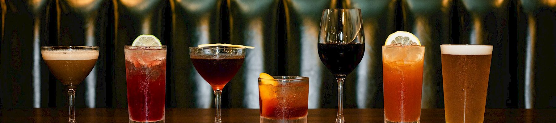 A row of seven varied cocktails on a bar counter, each glass filled with different colors, from light amber to dark red, with a green tufted backdrop.