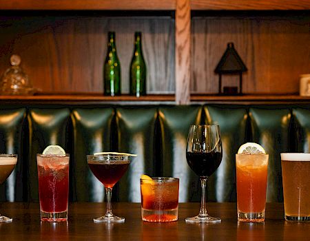 A row of seven varied cocktails on a bar counter, each glass filled with different colors, from light amber to dark red, with a green tufted backdrop.