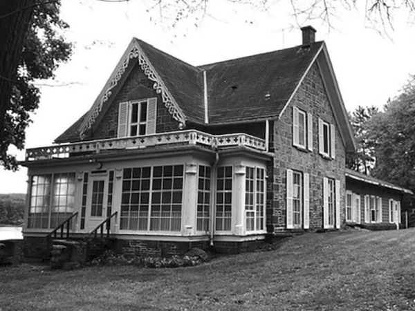 A black-and-white photo of a large wooden house with a wraparound porch, gabled roof, and many windows, set on a grassy yard.