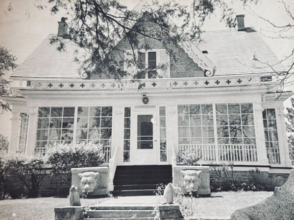 A vintage house with a symmetrical white porch, front steps, large windows, and decorative gables; framed by trees and shrubs, sunny yard.