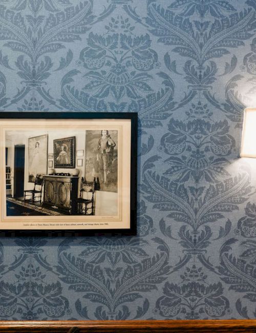 A blue damask-patterned wall with a framed black-and-white photograph and a wall sconce on the right, casting warm light.