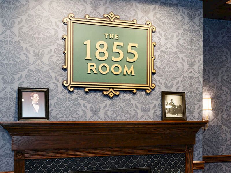 A cozy dining room by a lit fireplace, ornate blue wallpaper, a wooden mantel, and a sign reading &ldquo;The 1855 Room&rdquo; above the hearth, set for a meal.