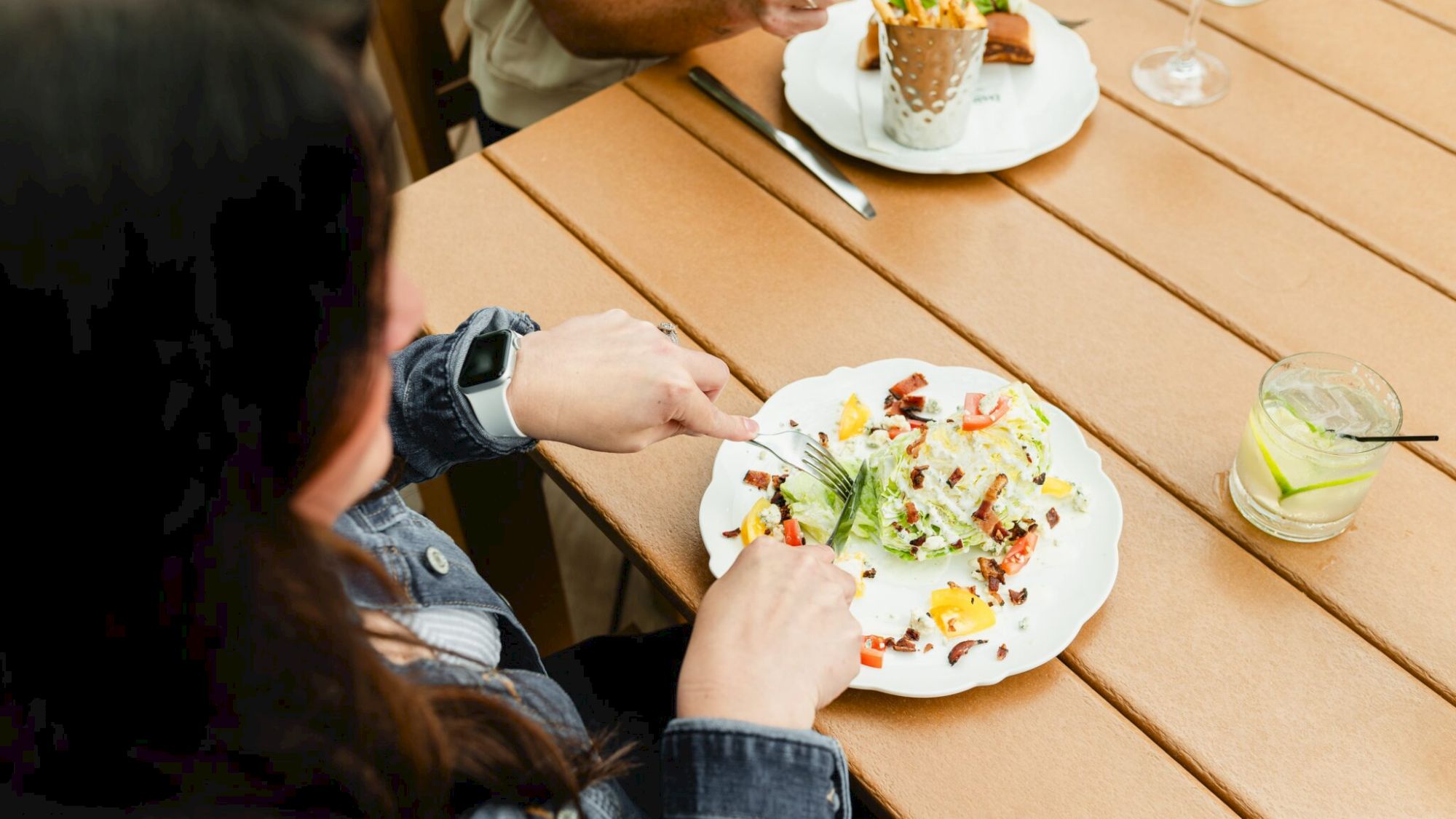 Two people are eating: a plate with colorful food and a drink on a wooden table. There&rsquo;s a dessert and another plate in the background.