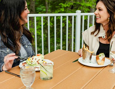 Two women sit at a wooden table outdoors, sharing drinks and salads with plates of food and smiling at each other.