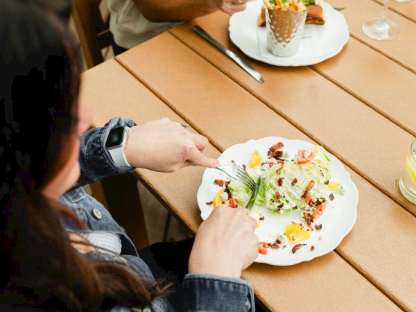 Two people share a picnic-style meal on a wooden table: plates with colorful food, a drink, and a dessert cone in the background, captured casually.