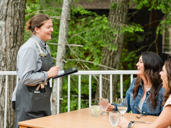 A waitress in a denim uniform takes a group order at an outdoor table, smiling as two friends chat and share drinks on a sunny day.