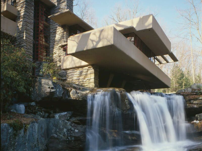 A modern house with a dramatic cantilevered balcony sits above a cascading waterfall and rocky ledges, blending architecture with nature.